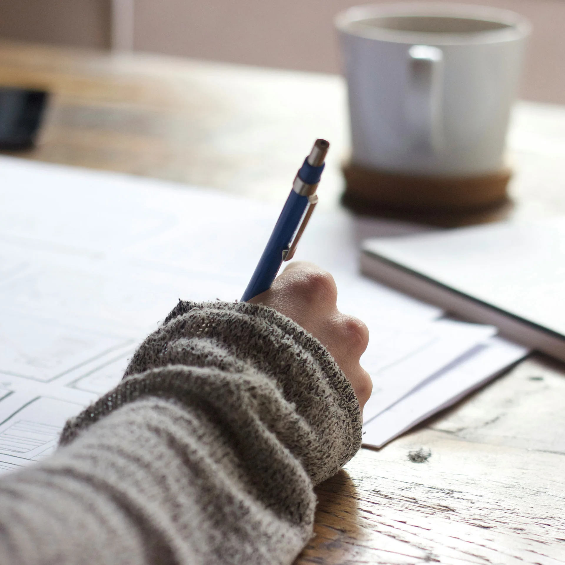 Hand som håller i penna och skriver med kaffekopp i bakgrunden